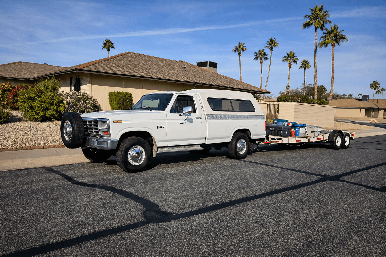 White Ford F-250 with camper shell towing a loaded flatbed trailer on a sunny residential street.